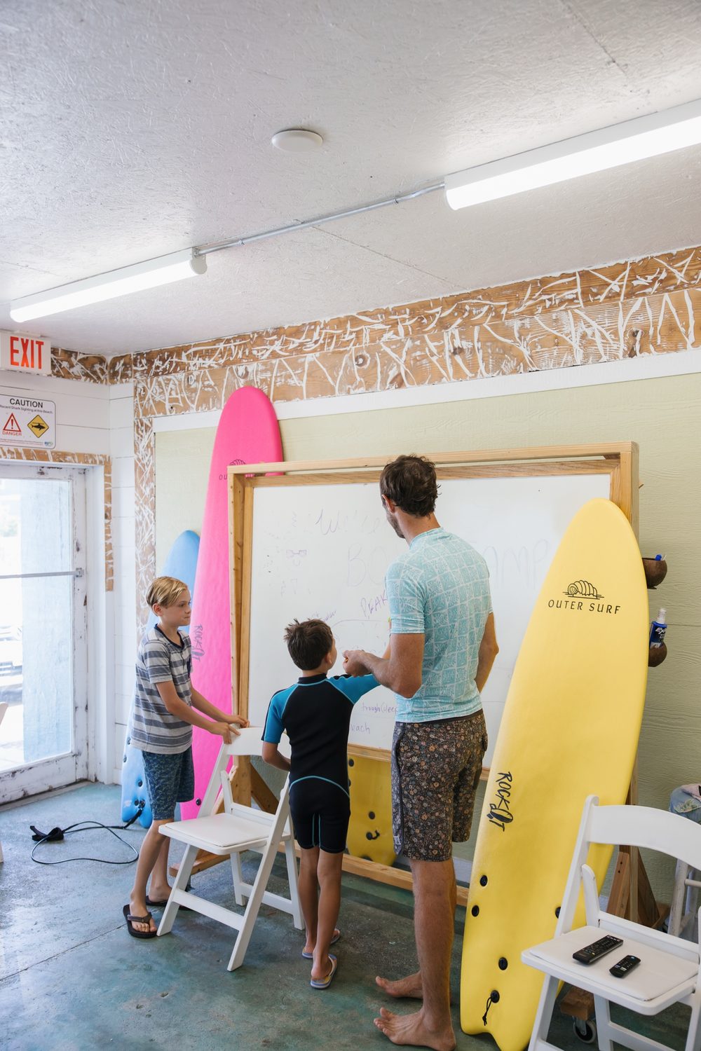 An instructor teaching kids about waves and sandbars at a whiteboard surrounded by surfboards