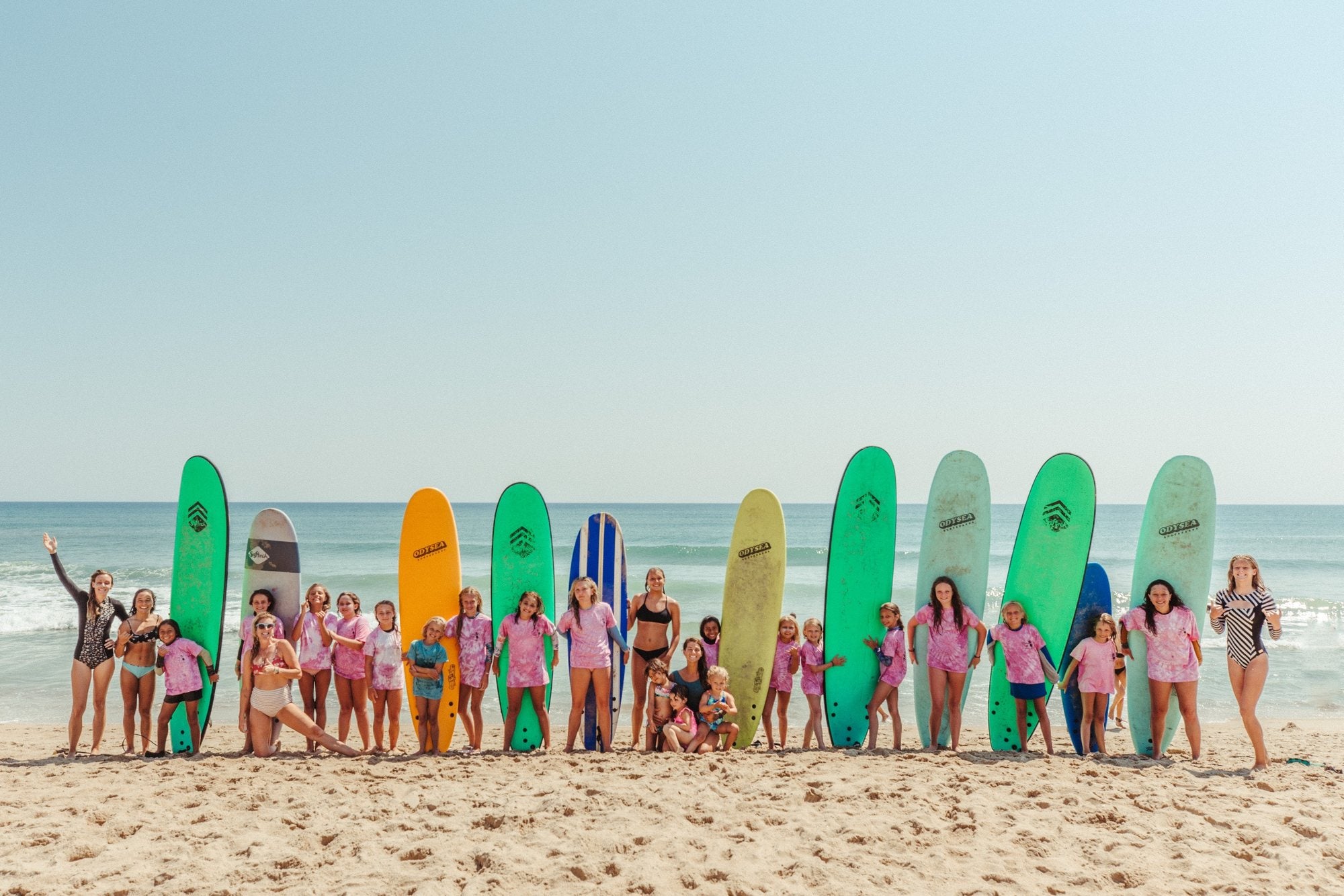 A long line of Girl Grom campers and instructors in pink tie-dye shirts standing with colorful soft-top boards on the beach