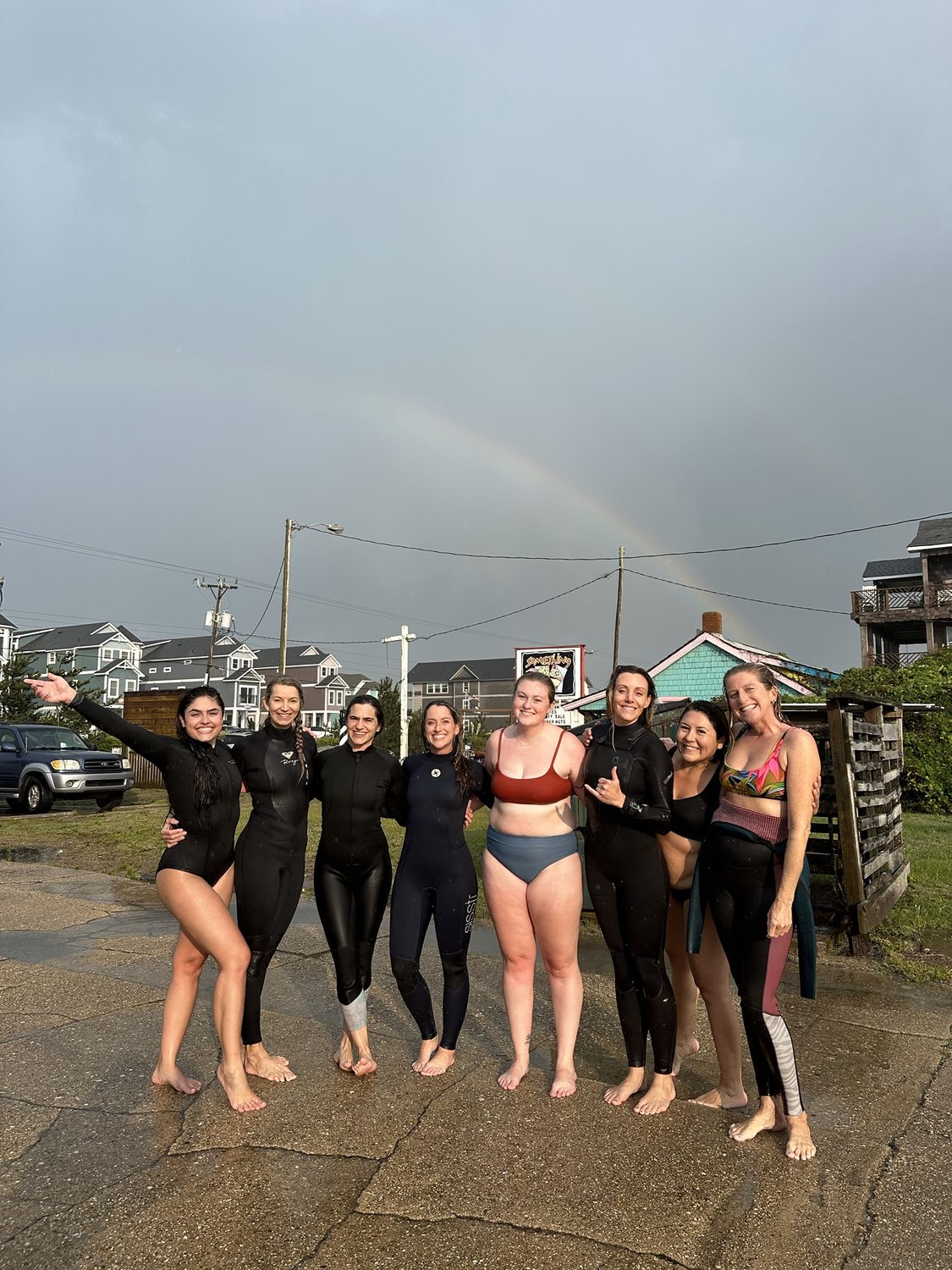 A women's surf crew in wetsuits smiling under a rainbow after a session