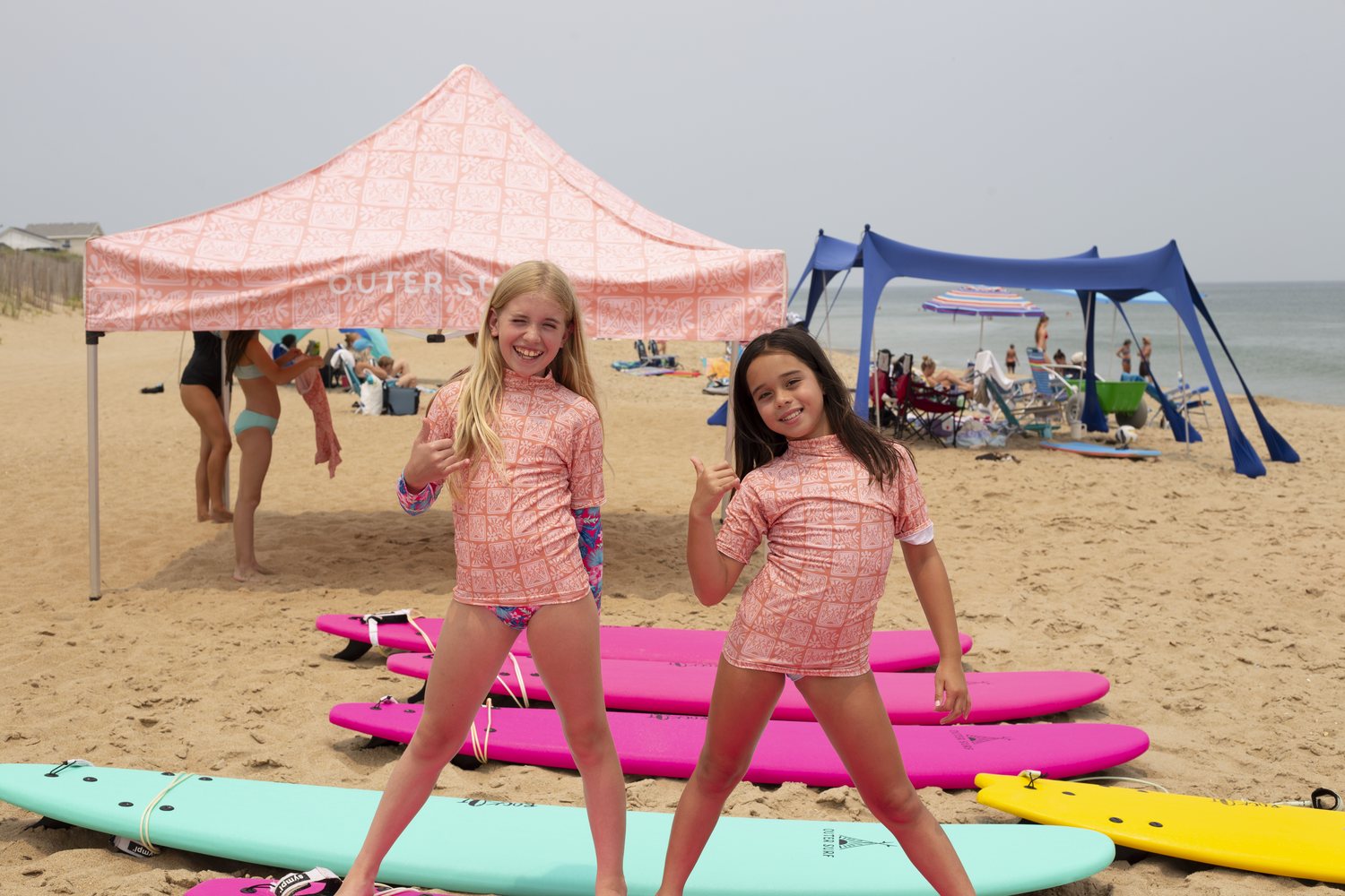Two Girl Grom campers throwing shakas in front of the Outer Surf tent and pink boards